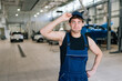 © dikushin - Confused car mechanic male in overalls feeling puzzled holding visor of his cap, looking at camera with serious expression. Technician man doing car maintenance in auto repair workshop.