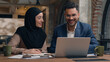 © Yuliia - Two business colleagues co-workers talk discuss corporate work at office cafe multiracial male female employees Indian man muslim woman in hijab diverse businesswoman businessman write tasks laptop