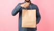 © Lola - Close up male holding in hand brown clear empty blank craft paper bag for takeaway isolated on pastel pink background. Packaging template mock up. Delivery service concept. Copy space advertising ar
