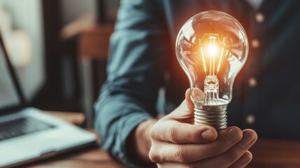 Poster - A person holds a glowing light bulb, symbolizing ideas and innovation, with a laptop visible in the background.