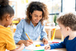 © Inga Bulgakova - A speech therapist in a denim shirt helps two young children with language exercises at a table, using colorful stationery in a bright, engaging classroom environment.