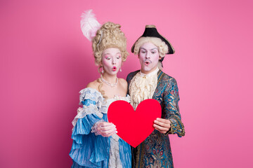 Wall Mural - Playful couple in vintage costumes holding a heart-shaped card against a pink background, capturing a humorous and romantic moment