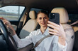 © SHOTPRIME STUDIO - Woman in car driver's seat, taking a selfie by holding up her phone to the camera, casual and candid moment, urban setting