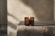 © Thedinindu - A lit brown candle in a glass jar sits on a rough, textured stone surface, bathed in soft sunlight.