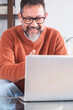© simona - Middle-aged man wearing glasses and casual clothing, seated comfortably on a sofa at home while working remotely on a laptop, seamlessly combining freelance tasks with personal online leisure