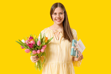 Wall Mural - Beautiful young pregnant woman with bouquet of tulips, greeting card and gift box on yellow background. International Women's Day