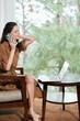 © SHOTPRIME STUDIO - Young woman in a brown dress sitting on a chair and talking on a smartphone while using a laptop at home, surrounded by greenery