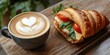 © Rasim - Cappuccino paired with a delicious croissant filled with cheese and tomato creates a stylish view. Enjoy the latte art heart design in the coffee cup beside the croissant sandwich on the table.
