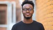 © Wanida - Confident young man smiling while standing against an urban brick wall, exuding positivity and approachability with modern eyeglasses and casual attire.