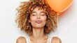 © Katewaree - Young woman with curly hair expressing happiness while holding an orange balloon against a light background, radiating positivity and carefree vibes