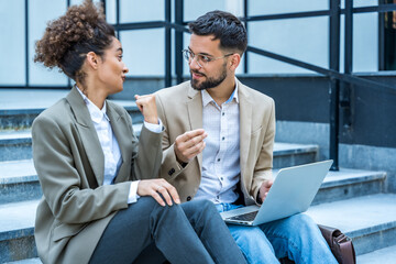 Two business professionals standing outside a modern office building, engaged in a discussion. Businessman and businesswoman sitting on stairs in front of office building, working on laptop computer.