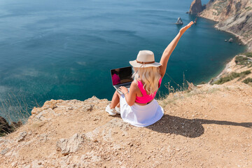 Wall Mural - A woman is sitting on a rocky beach with a laptop in front of her. She is wearing a pink top and a straw hat. She is waving her hand in the air, possibly to celebrate or to show excitement.