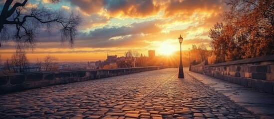 Poster - Scenic cobblestone road leading to city center under dramatic sunset sky with trees and lamppost creating a tranquil atmosphere