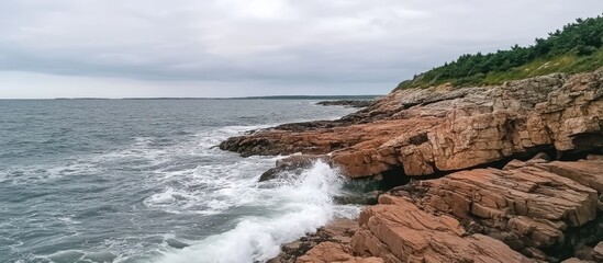Poster - Waves crashing on rocky coastline under a cloudy sky creating a serene coastal landscape with lush greenery in the background