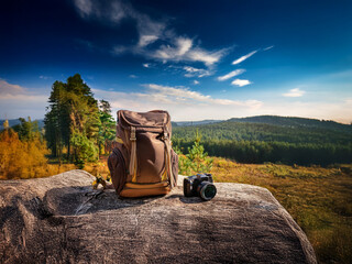 Poster - A brown backpack and camera rest on a rock overlooking a picturesque autumn landscape.  Sunlight bathes the scene, promising adventure.