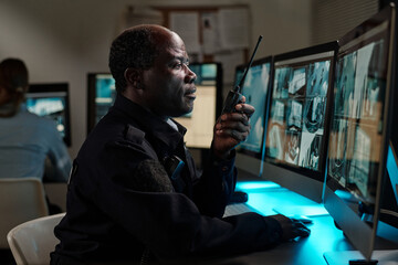 Poster - Mature African American male officer sitting in front of computer monitor and looking at screen while speaking on walkie-talkie