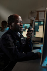 Poster - Mature African American security guard in black uniform looking at desktop computer screen while speaking on walkie-talkie