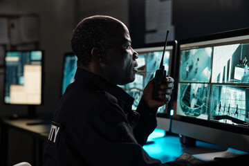 Poster - Side view of mature African American male guard with walkie-talkie in hand sitting in front of desktop computer monitors