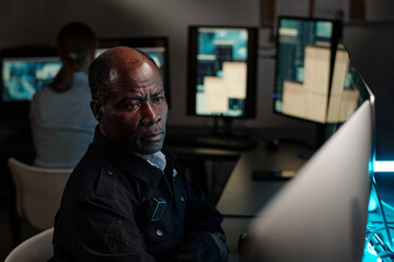 Poster - Serious mature security guard sitting by workplace in observation room and looking at one of desktop computer screens