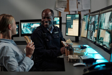Poster - Smiling mature male security guard with sandwich looking at young female colleague during chat at lunch break