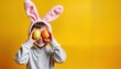 © miss irine - Adorable child wears Easter bunny ears, holds colorful eggs in front of eyes. Happy kid enjoying Easter holiday. Child dressed in light gray hoodie, smiling broadly against bright yellow background.