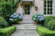 © gamespirit - A front door of an old brick house with blue hydrangeas in large planters on each side. A cobblestone walkway leads to the door, surrounded by tall hedges and flowers. A pretty, symmetrical scene, in