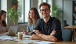 © kotlyarn - A cheerful young man with glasses sits confidently at a wooden table in a bright, modern office, surrounded by two women engaged in work. The atmosphere is vibrant and collaborative, showcasing a
