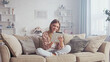 © MIKHAIL - Young woman sitting comfortably on a sofa in her living room, using a digital tablet with a cheerful expression, enjoying modern technology in a cozy home environment
