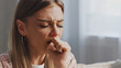 © MIKHAIL - Young woman coughing and feeling unwell, eyes closed and hand covering her mouth, sitting on a sofa in her living room, experiencing discomfort and fatigue from illness