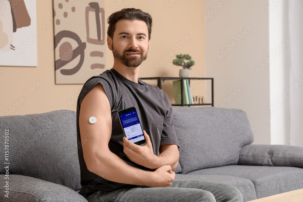 Diabetic man with glucose sensor using mobile phone for measuring blood sugar level on sofa at home