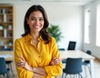 © Viktor - Confident Latin American businesswoman stands in modern office. Smiling woman looks directly at camera with arms crossed. Wears bright yellow shirt. Successful, pro person in corporate environment.