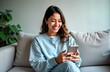 © Viktor - Happy young Hispanic woman uses smartphone at home. Smiling female teen scrolls social media shops online. Relaxed on comfy couch. Casual wear. Modern lifestyle. Digital services. E-commerce. Mobile
