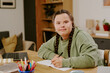 © AnnaStills - Medium portrait of confident young woman with disability sitting at wooden desk in living room, looking at camera while writing in notebook
