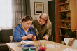 © AnnaStills - Senior woman standing at table in living room, teaching her daughter with Down syndrome how to embroider