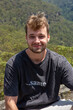 © Keely Anne - man stands confidently on a rocky ledge overlooking a vast, green valley under a clear blue sky. Dressed casually, he blends into the serene natural setting, radiating calm and connection with nature