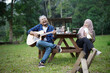 © Fajar - Indonesian man and young Indonesian woman in hijab playing guitar and singing together at picnic table while going camping in forest cabin, travel concept.