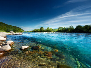 Poster - Crystal-clear river flows gently through a serene landscape,  under a vibrant blue sky.  Large rocks line the shallows, and lush green trees border the banks.