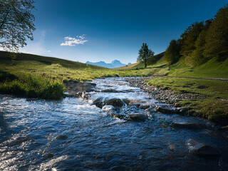 Poster - Serene stream flows through a verdant valley, nestled between rolling hills and majestic mountains under a clear blue sky.  Sunlight illuminates the tranquil scene.