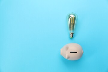 Wall Mural - Piggy bank and lightbulb on light blue background, flat lay with space for text. Energy saving concept