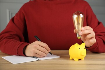 Wall Mural - Man holding light bulb above piggy bank while taking notes at wooden table, closeup. Energy saving concept