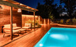 © Mariakray - Modern outdoor lounge area with wooden pergola and swimming pool at night. Luxury backyard illuminated with soft lighting