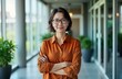 © Maryna - Mature Asian businesswoman stands confidently in office building hallway. Wears glasses, orange shirt with arms crossed. Looks directly at camera. Indoor shot. Successful professional. Powerful