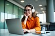 © Maryna - Young woman working on laptop inside modern office. Wears stylish orange shirt, headphones. Woman smiling, listening to online podcast audio course. Sits at workplace in contemporary office. Enjoying