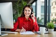 © Maryna - Young woman in red blazer, shirt wearing headphones with microphone sits at desk in modern office. Happily looks at camera, takes notes on documents in front of. Bright, light office space with