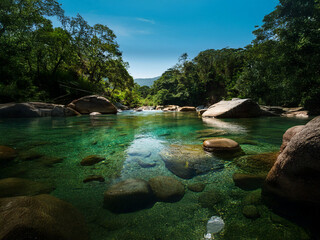 Poster - Serene river scene, crystal-clear water reveals smooth river rocks on the bed. Lush green vegetation frames the tranquil waterway under a vibrant blue sky.  A glimpse of distant hills adds depth to th