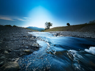 Poster - Serene stream flows through a rocky landscape,  under a vibrant blue sky. A lone tree stands on a small hill overlooking the tranquil scene.  Sunlight brightens the horizon.