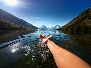 Poster - A hand reaches into a pristine mountain lake, splashing water.  Sunlight glistens on the calm surface, reflecting the majestic peaks.  Serene, idyllic scene.