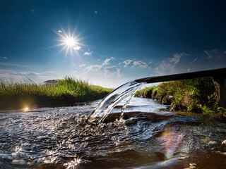 Poster - Clear water flows from a pipe into a tranquil stream under a bright sun. Lush greenery borders the waterway, creating a serene, natural scene.
