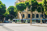 A modern tram travels along the street in Rio de Janeiro, surrounded by lush green trees and historic buildings.