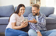 © Studio Romantic - Joyful Caucasian family couple sitting on floor near sofa, using mobile phone together for online entertainment at home. Happy husband and wife laughing while surfing internet or watching funny videos